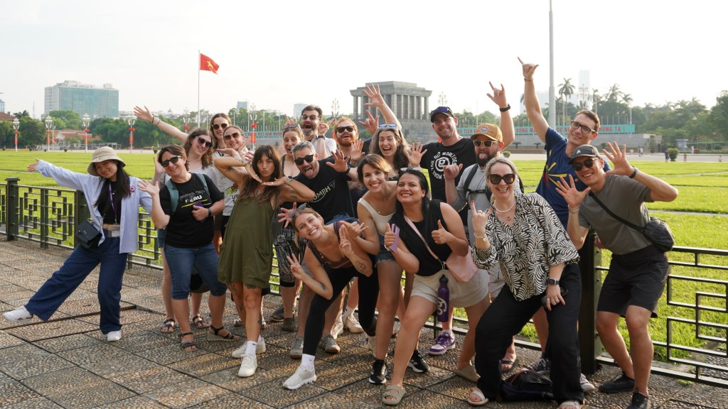 A diverse group of people, smiling and posing joyfully together in a park setting, with a large flag of Vietnam in the background and a historic building visible.